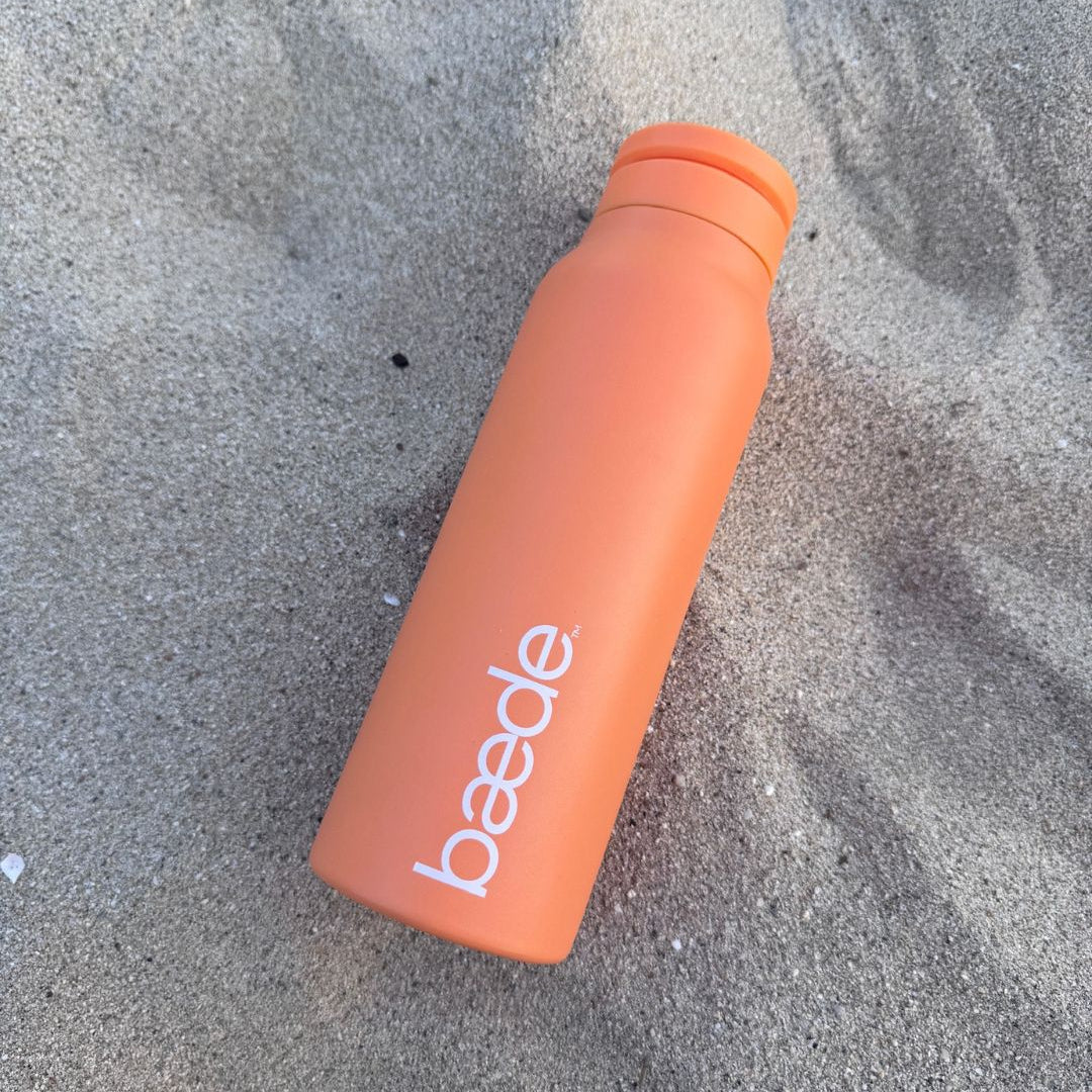 Sunset orange insulated water bottle with the brand name 'baede' printed in white, lying diagonally on a textured gray sandy surface.