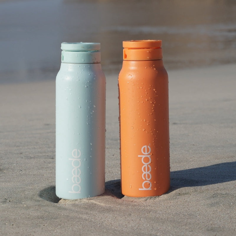 Two matte insulated water bottles from Beade standing in sand on a beach. One bottle is a mint icy blue and the other is a sunset orange, both with condensation droplets and the Beade logo printed vertically.