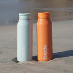 Two matte insulated water bottles from Beade standing in sand on a beach. One bottle is a mint icy blue and the other is a sunset orange, both with condensation droplets and the Beade logo printed vertically.