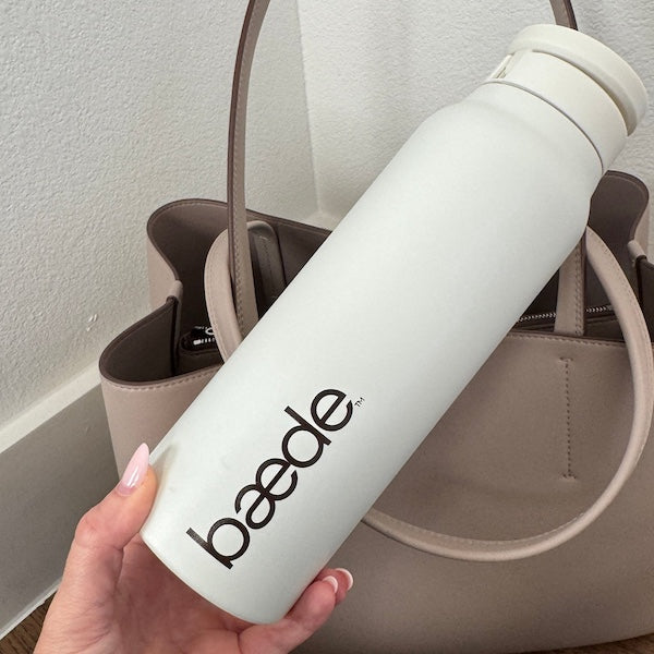 A hand holding a cream insulated water bottle with the brand name 'baede' printed in dark brown, positioned in front of a taupe tote bag on a white wall and wood floor.